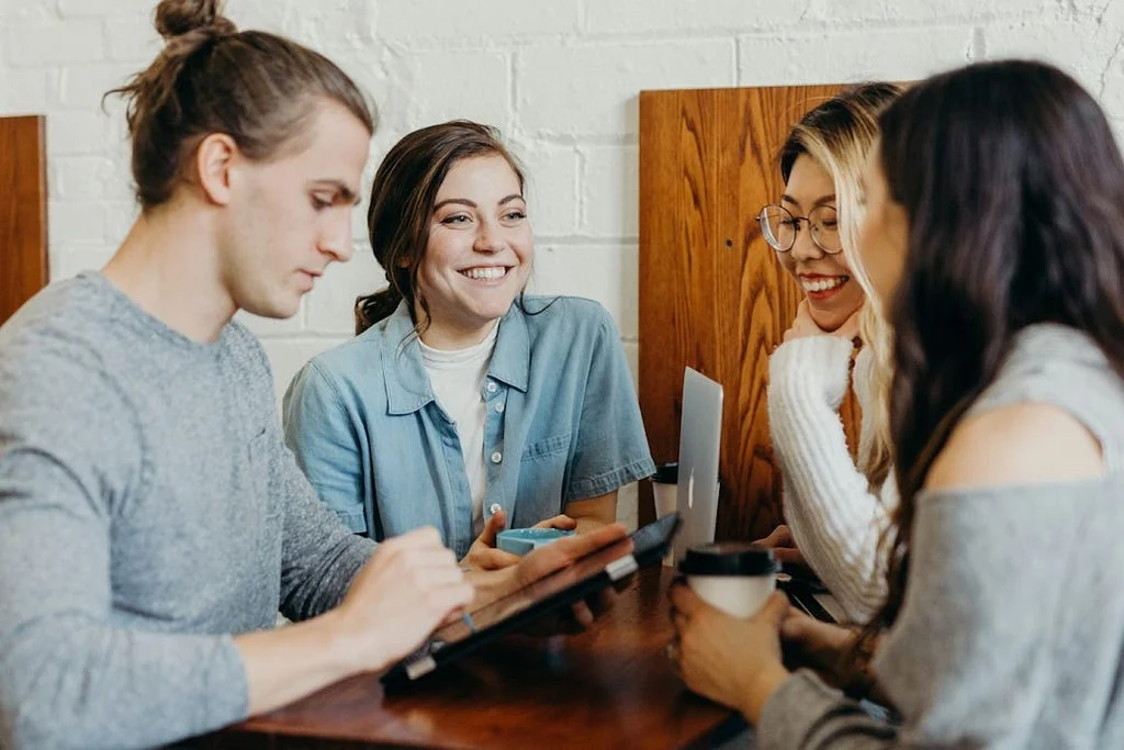 Four young colleagues gathered around a table, smiling, using a laptop and tablet with coffee cups nearby.