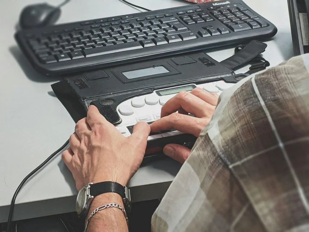 Close-up of two hands typing on a braille keyboard/note-taking device positioned in front of a standard keyboard.