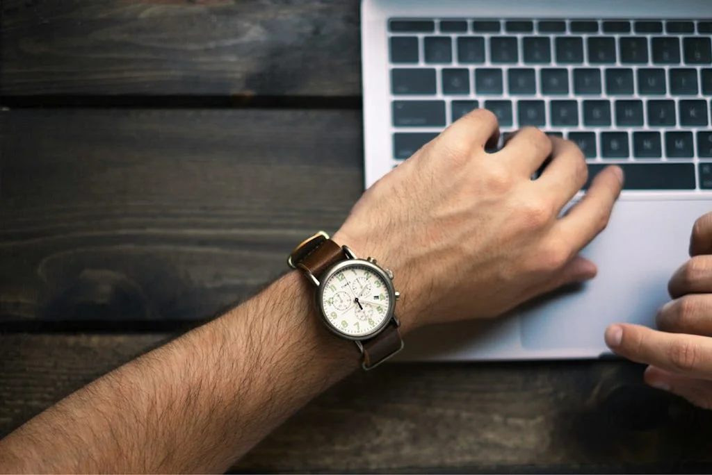 A person's wrist wearing a watch while typing on a laptop keyboard, set on a dark wood table.