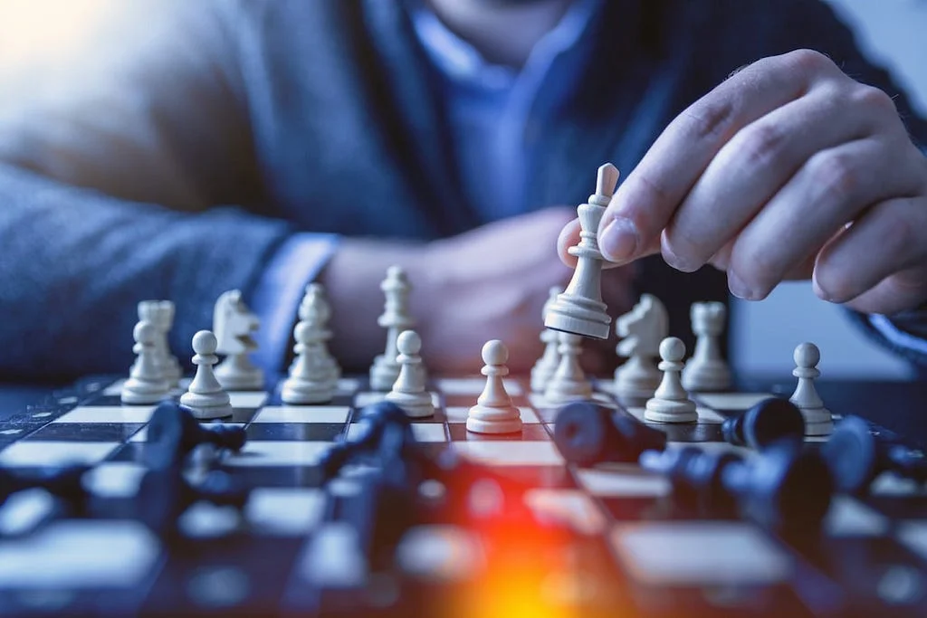 A hand moving a white king chess piece across a chessboard, with fallen black pieces in the foreground.