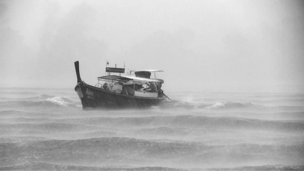 A black-and-white photo of a small longtail boat navigating rough, stormy seas in heavy rain and fog.