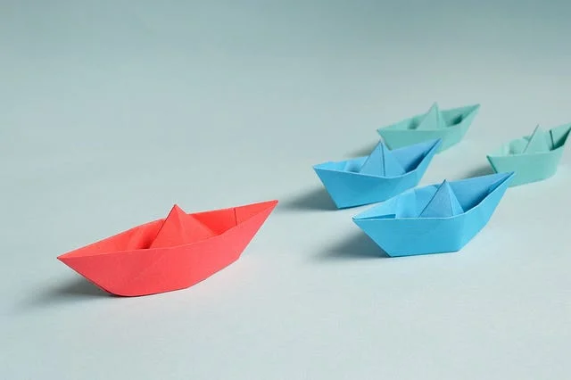 A red paper boat leading ahead of a group of blue and teal paper boats on a plain background.