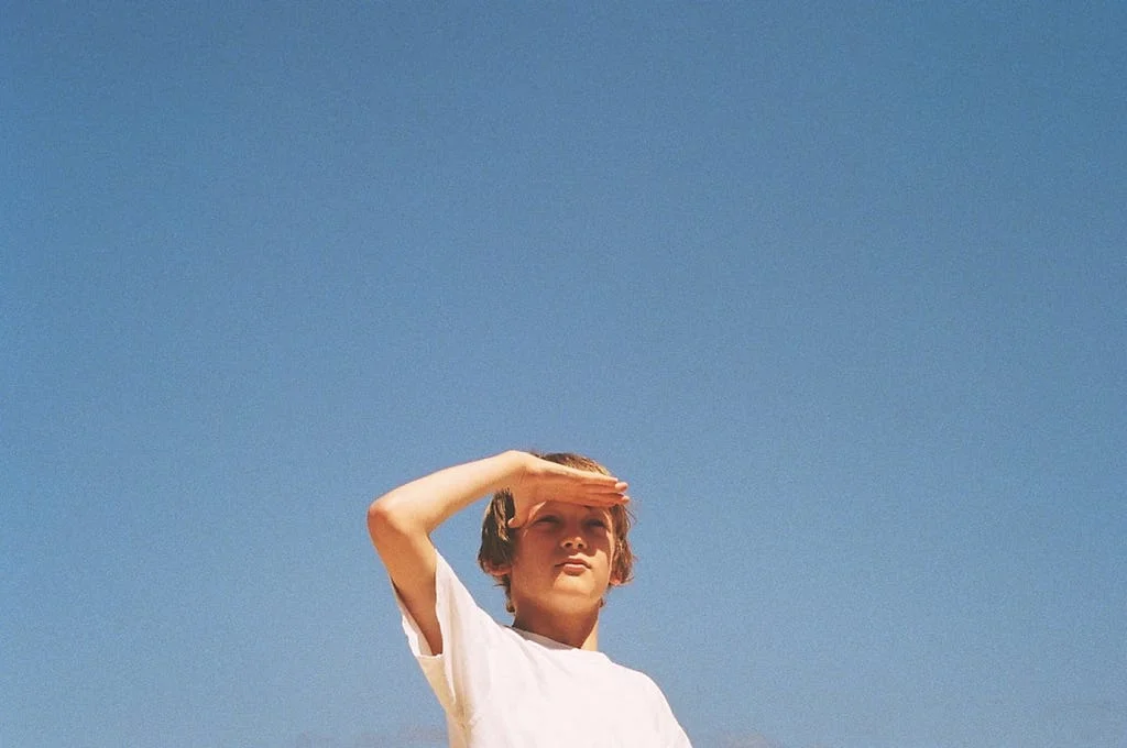 A young person outdoors shielding their eyes from bright sunlight against a clear blue sky, squinting upward.