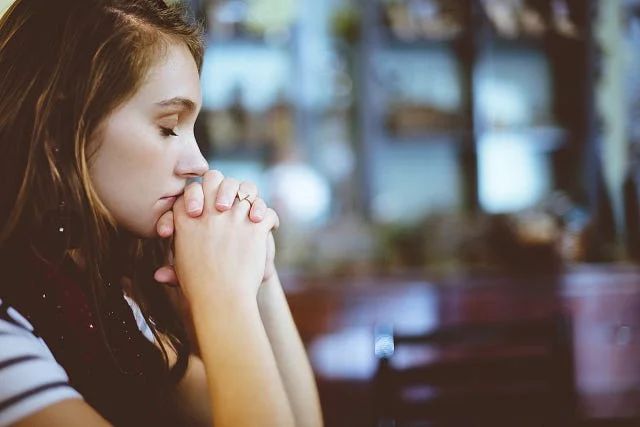 A young woman sitting with her hands clasped in front of her face, eyes closed, in a quiet indoor space.